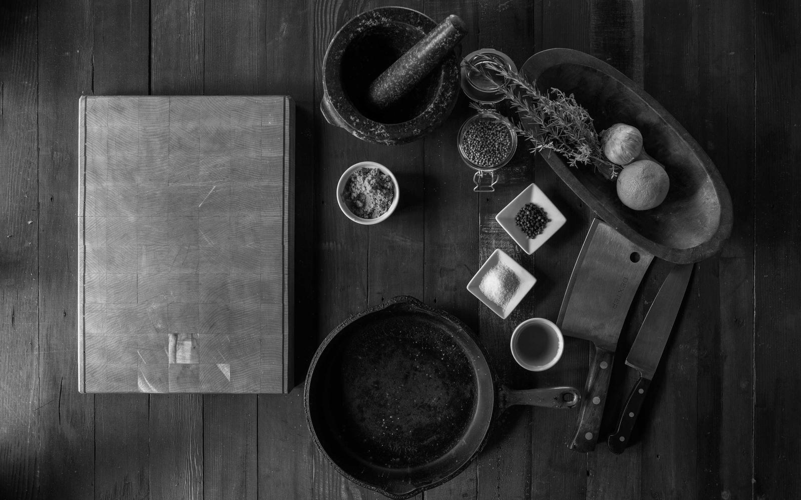 A dark kitchen worktop with a chopping board, pan and knives seen from above